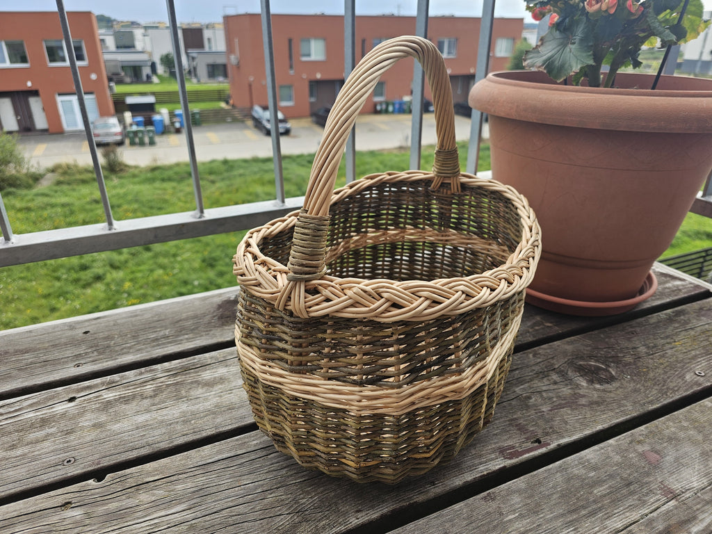 Oval handwoven willow market basket with handle and braided rim on a wooden balcony table beside a terracotta flower pot.