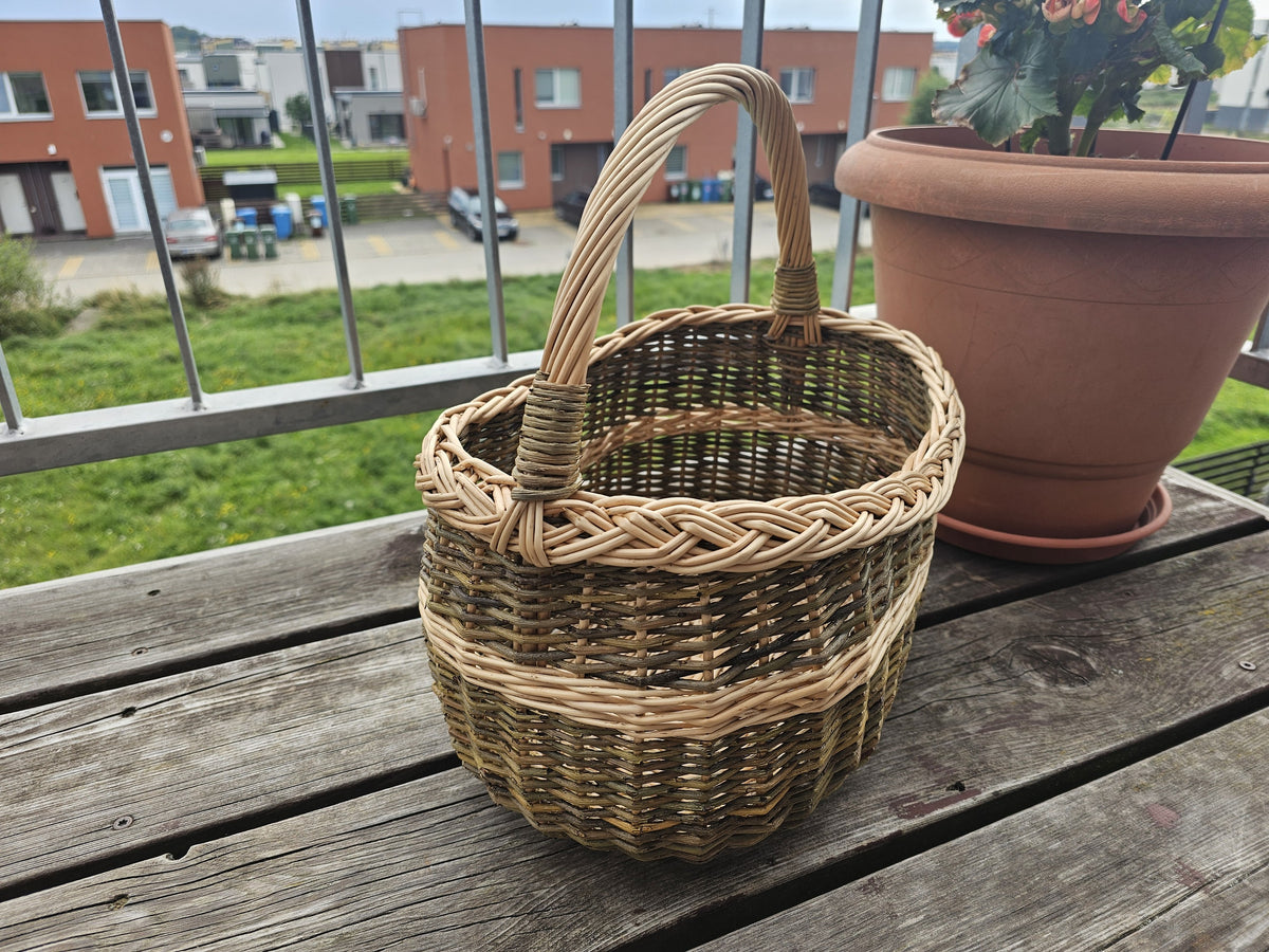 Oval handwoven willow market basket with handle and braided rim on a wooden balcony table beside a terracotta flower pot.