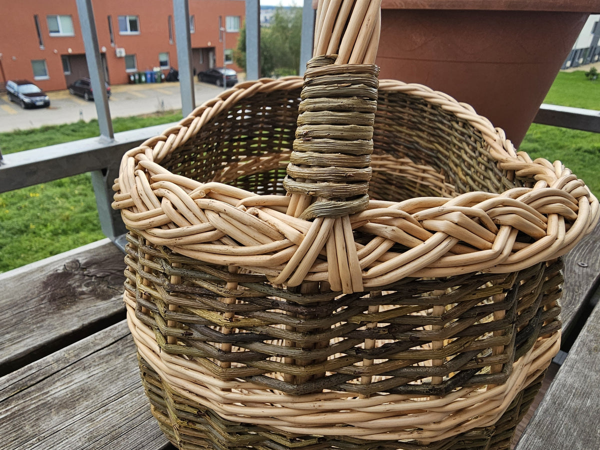 Close-up of the braided rim and wrapped handle joint of an oval handwoven willow shopping basket on a balcony bench.