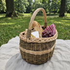 Handwoven oval picnic and market basket with a braided rim holding a baguette, cheese, grapes and a checked napkin on a linen blanket in a park.