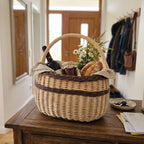 Willow market basket with handle carrying a bottle of wine, a loaf of bread, a jar and field flowers on a wooden hallway dresser.