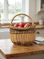 Wicker harvest basket with a tall handle filled with red apples on a wooden kitchen counter near a bright window