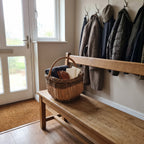 Handwoven willow basket with a tall handle holding gloves and a scarf on a wooden hallway bench beside hanging coats