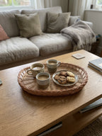 Oval wicker bread tray with a braided rim on a living room coffee table, serving three cups of coffee and a plate of cookies beside the sofa