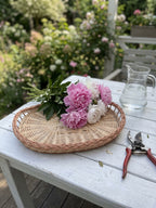 Handwoven willow serving platter with a braided rim holding a bunch of peonies on a white garden table next to a water jug and pruning shears