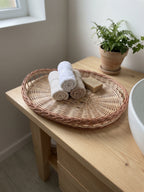 Oval willow tray used in a bathroom on a wooden vanity, holding rolled white towels and a bar of soap next to a potted fern