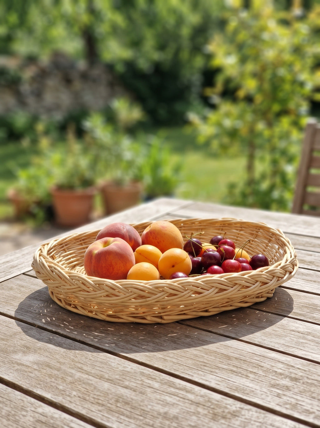 Handwoven oval wicker tray filled with peaches, apricots and cherries on a wooden garden table in summer sunlight