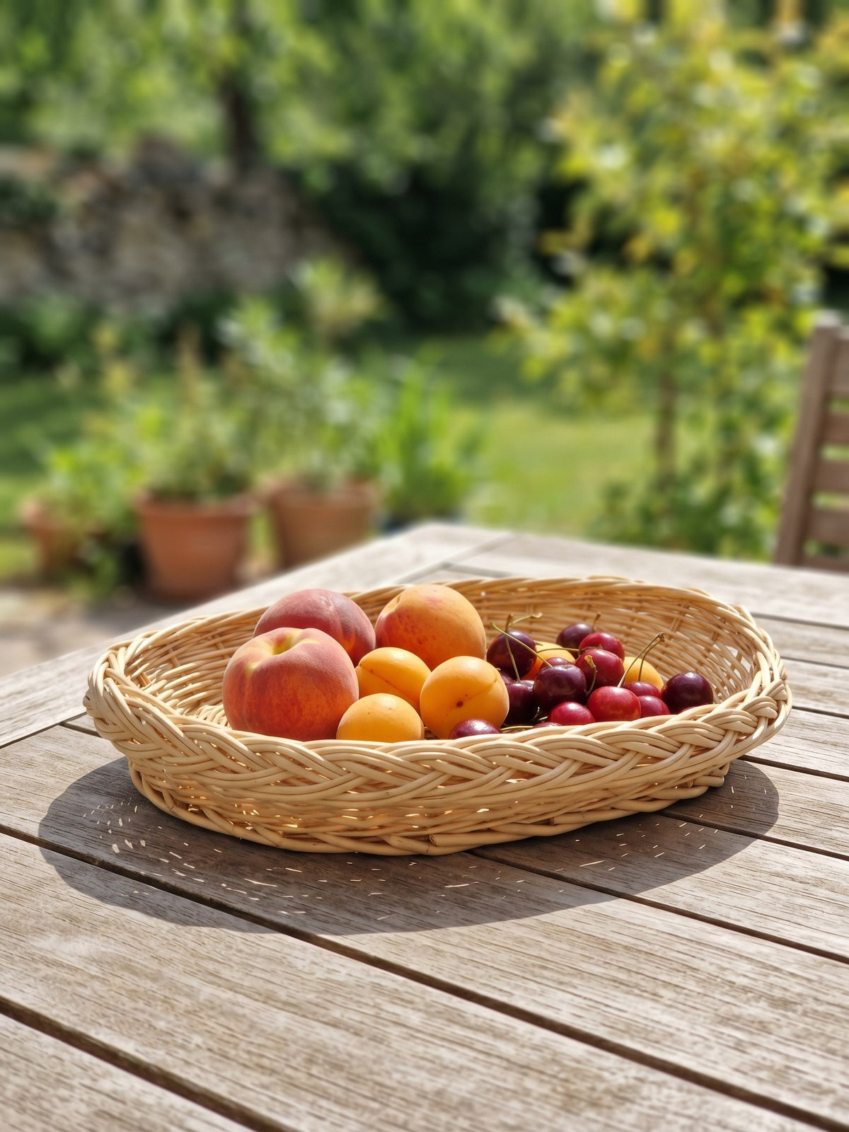Handwoven oval wicker tray filled with peaches, apricots and cherries on a wooden garden table in summer sunlight