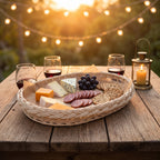 Wicker serving platter with a braided rim laid with cheese, salami, crackers and grapes on a rustic outdoor table with wine glasses and string lights
