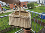 Hand lifting the willow garden basket by its two crossed handles on a balcony, showing how the handles meet at the top.