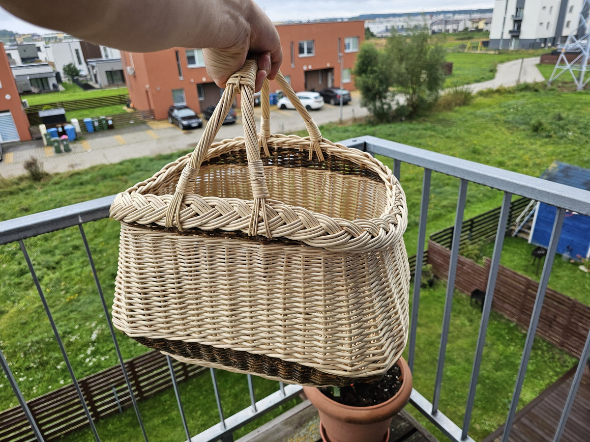 Hand lifting the willow garden basket by its two crossed handles on a balcony, showing how the handles meet at the top.