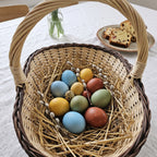 Handwoven oval Easter basket with handle holding dyed Easter eggs on straw with pussy willow twigs on a table set for a spring breakfast