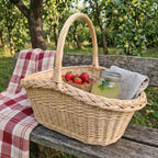 Oval willow gathering basket with handle holding strawberries, a jar of lemonade and a linen napkin on a garden bench with a checked blanket