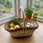 Oval willow kitchen basket with handle holding lemons, garlic and fresh thyme on a wooden windowsill next to a potted basil plant