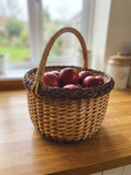 Handwoven round willow basket with a braided rim and smooth arched handle filled with red apples on a wooden kitchen counter