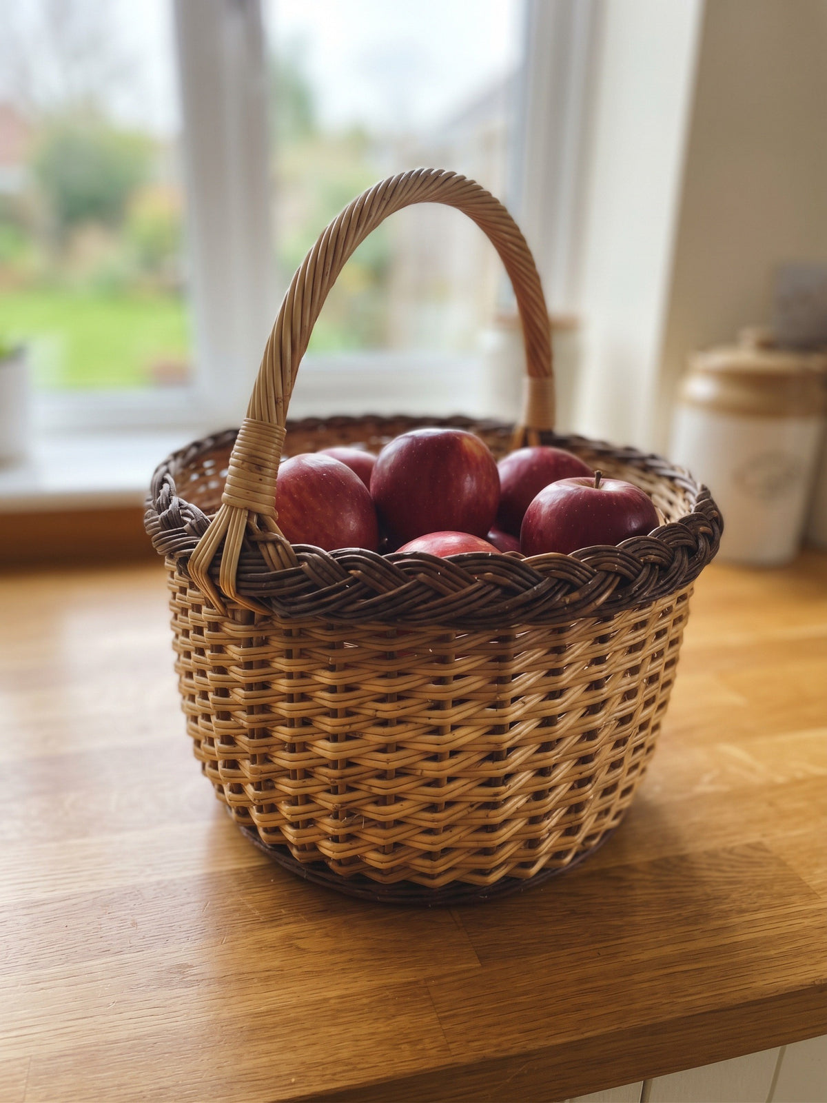 Handwoven round willow basket with a braided rim and smooth arched handle filled with red apples on a wooden kitchen counter