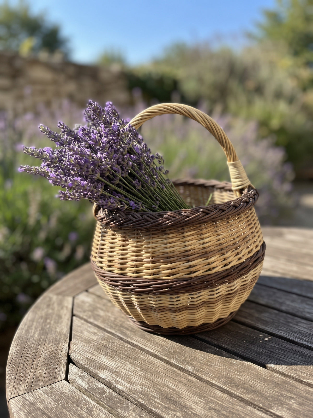 Small handwoven wicker flower basket with handle holding a bunch of lavender on a round wooden garden table