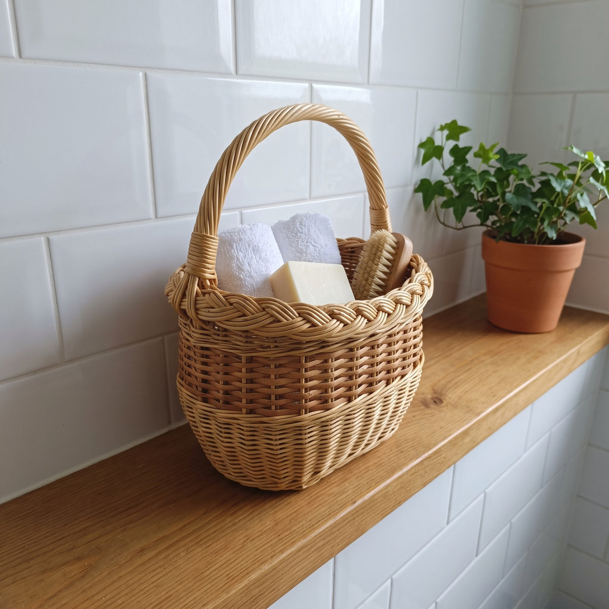 Small handwoven willow bathroom basket with handle holding towels, soap and a brush on a shelf against a tiled wall