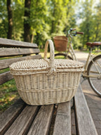 Small wicker picnic basket with a double lid and central handle, handwoven from willow, sitting on a park bench with a bicycle behind it.
