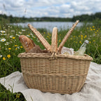 Handwoven willow picnic basket for two with both lids open, packed with a baguette, cheese and lemonade on a blanket by a lake meadow.