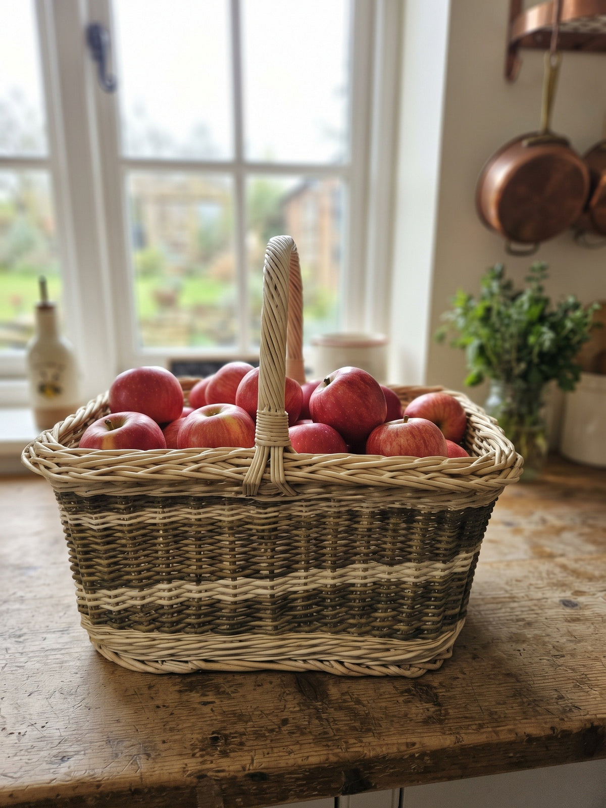 Rectangular willow vegetable harvest basket with a tall arched handle filled with red apples on a rustic kitchen worktop by the window