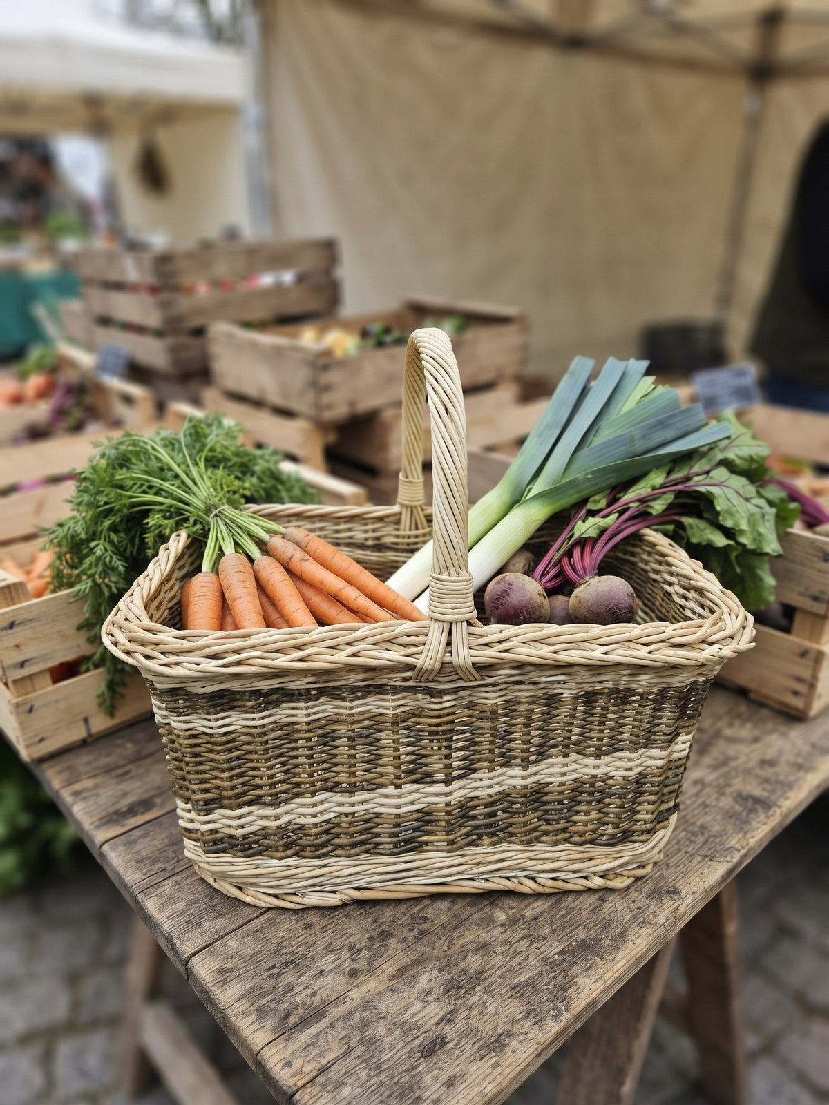 Rectangular wicker market carrier with handle holding carrots, leeks and beetroot on a wooden table at an outdoor farmers market