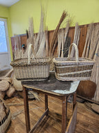 Two rectangular wicker shopping baskets with handles side by side on a wooden table, with bundles of willow rods behind them