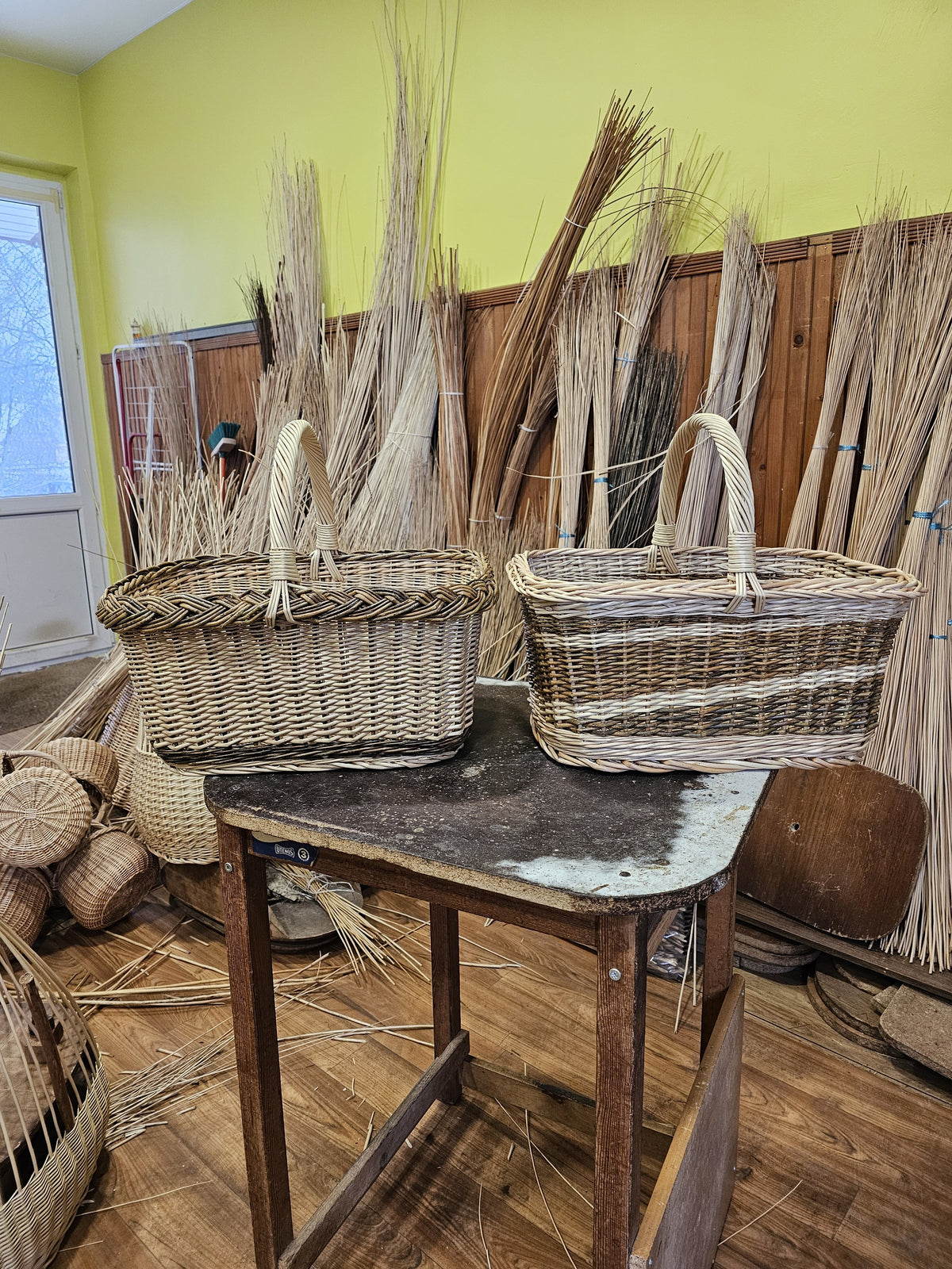 Two rectangular wicker shopping baskets with handles side by side on a wooden table, with bundles of willow rods behind them
