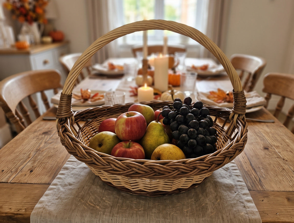 Round wicker garden trug with an arched handle holding apples, pears and grapes on a wooden dining table set for an autumn dinner