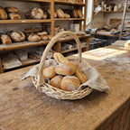 Round willow bread basket trug with handle holding crusty rolls on a linen cloth on a bakery counter with shelves of loaves