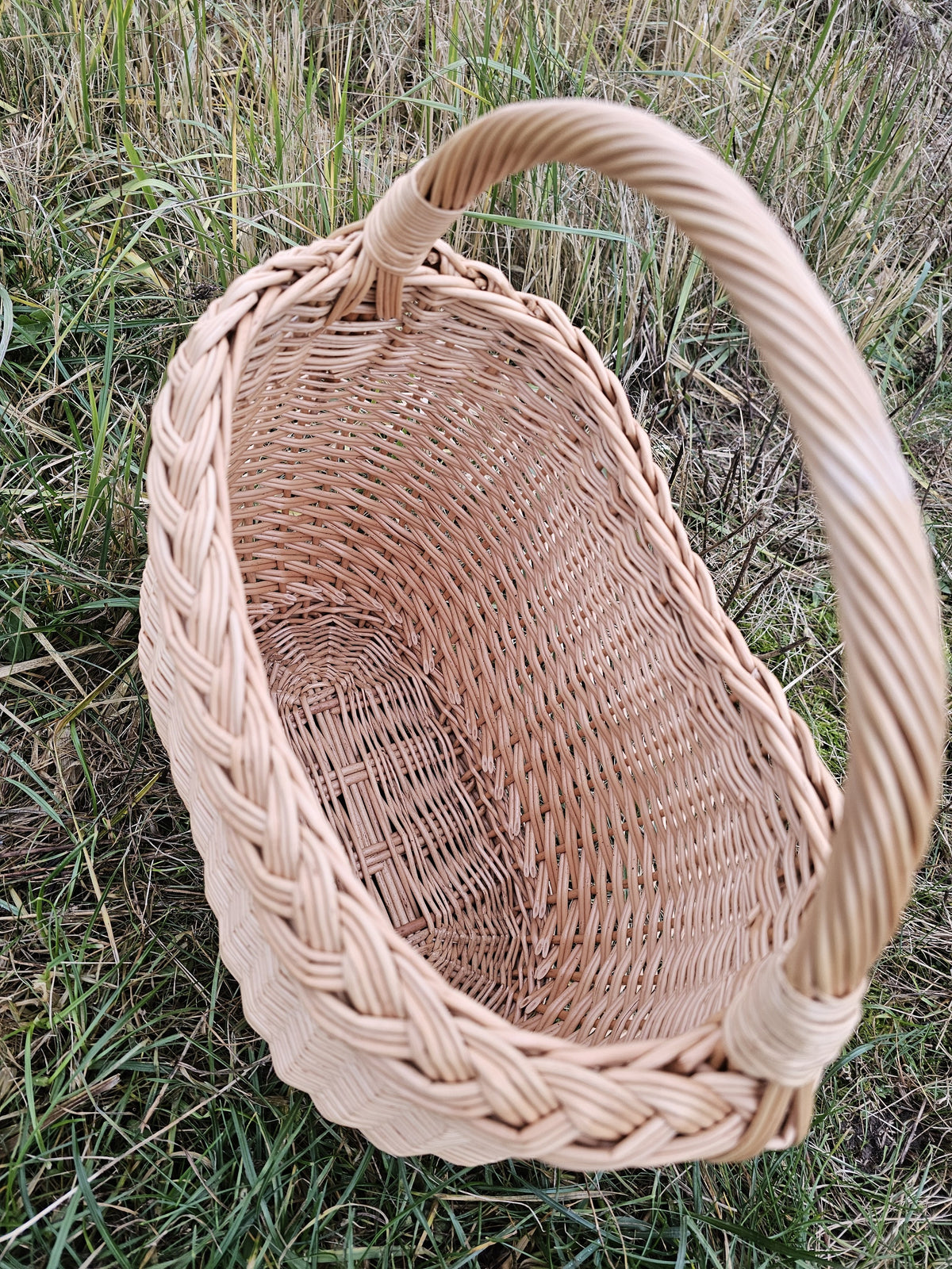 Close view into the empty willow basket in the grass, the thick plaited rim and generous curved handle showing the handmade construction.