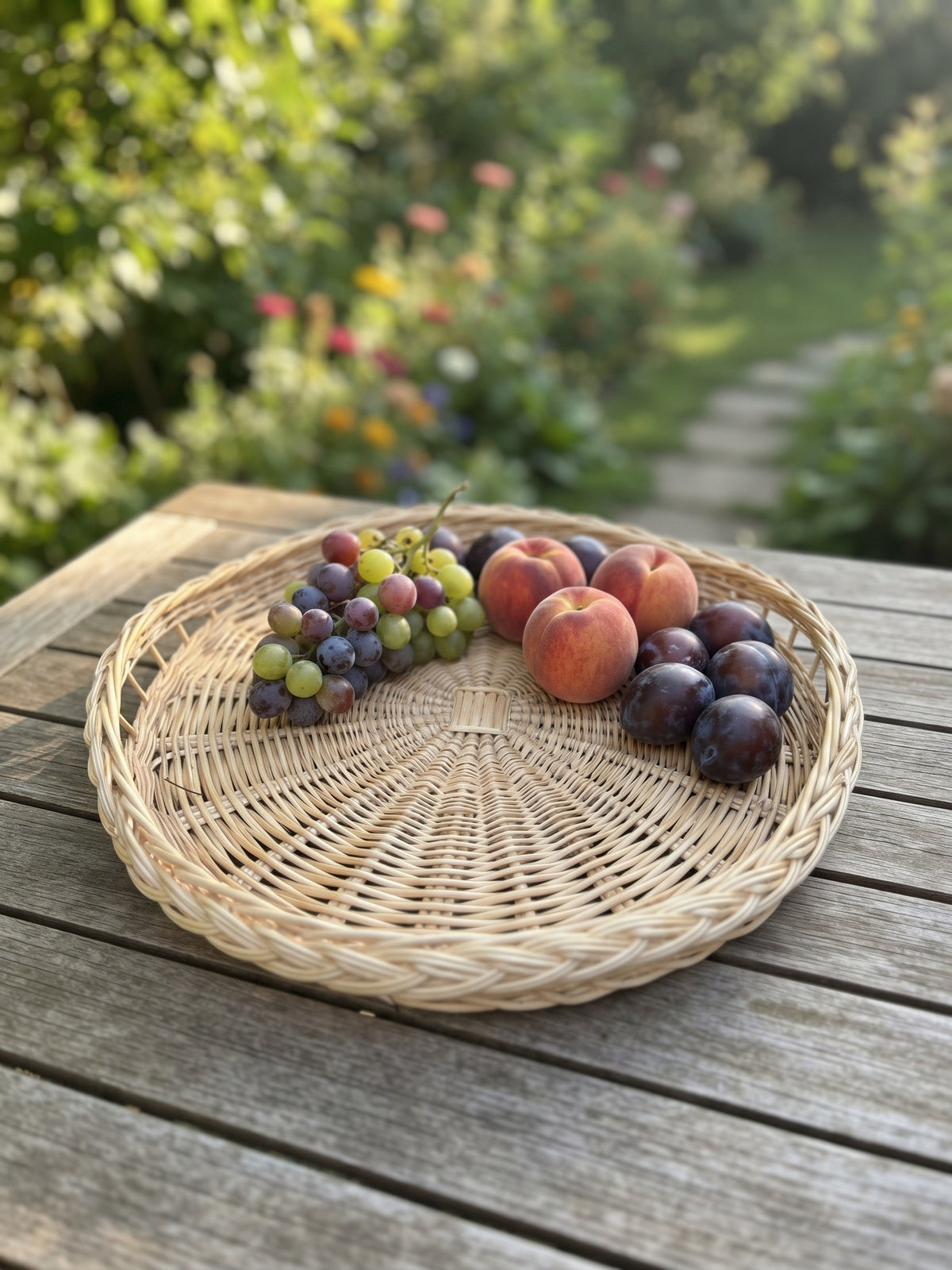 Round wicker serving tray with a braided rim on a weathered garden table, holding grapes, peaches and plums with a flowering cottage garden path behind.
