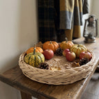Woven willow serving tray with a braided rim on a rustic wooden console, filled with small pumpkins, red apples and pine cones as autumn decor.