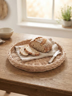 Round wicker bread tray on a kitchen table by the window, holding a sliced sourdough loaf on a linen napkin with a bread knife.