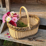 Ovaler Blumenkorb aus Weide mit Zopfrand und Bügelhenkel voller frischer Dahlien, daneben eine Gartenschere auf einer Holzstufe