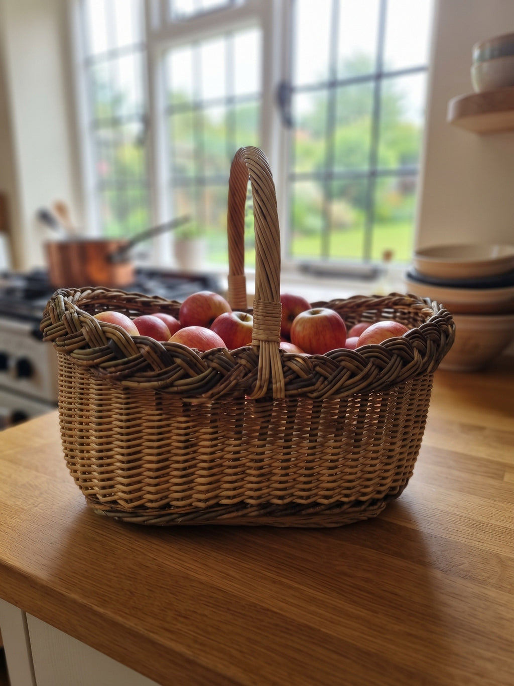 Oval wicker basket with a braided rim and handle filled with red apples on a wooden kitchen counter by a bright window