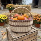 Oval wicker basket with a braided rim holding pumpkins and apples on a wooden crate at an autumn farm stand with hay bales and chrysanthemums