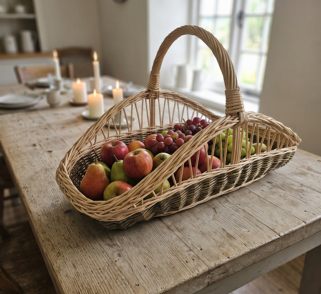Long willow trug with a tall handle filled with apples, pears and grapes on a farmhouse dining table with candles