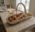 Long willow trug with a tall handle filled with apples, pears and grapes on a farmhouse dining table with candles