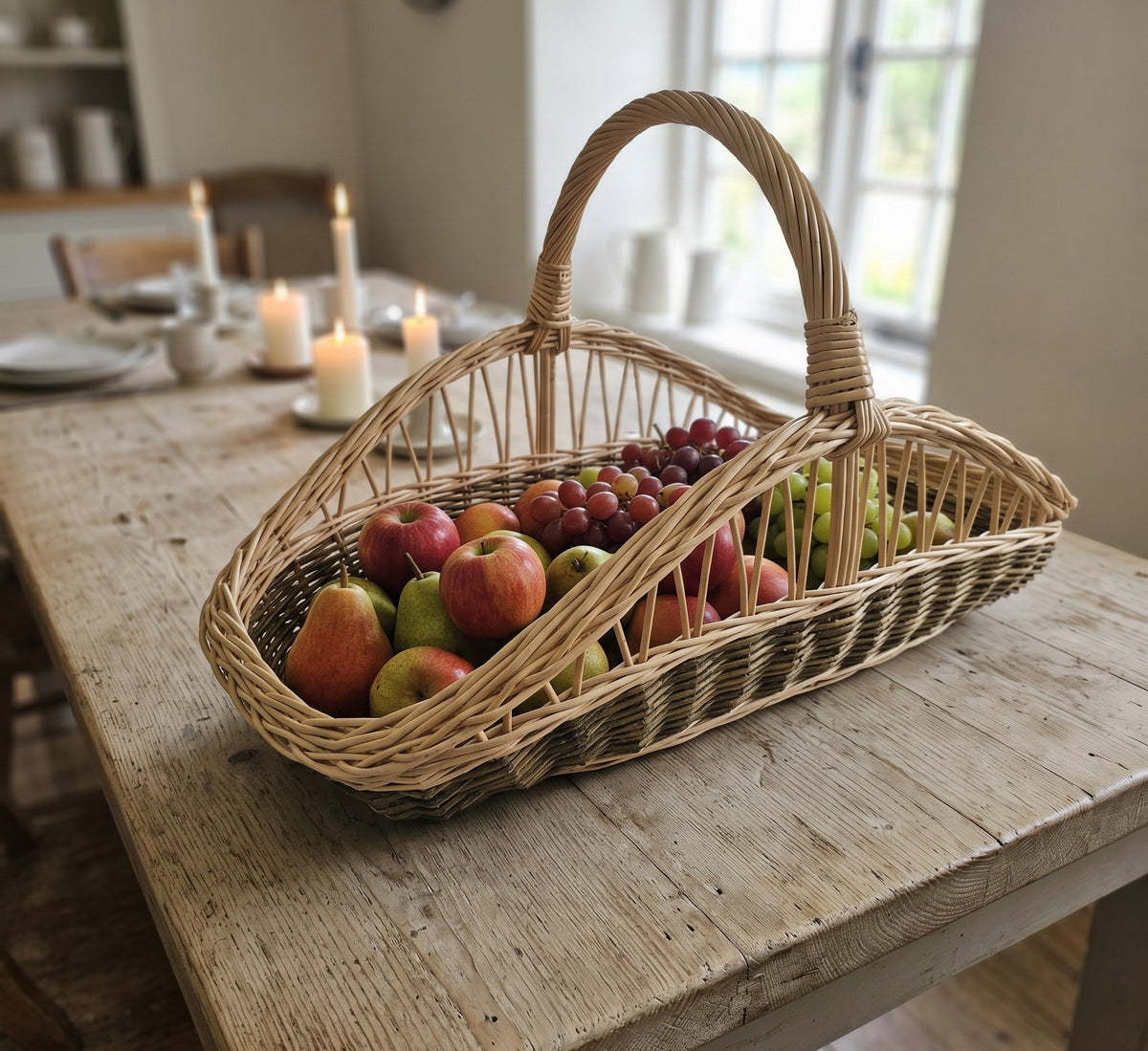 Long willow trug with a tall handle filled with apples, pears and grapes on a farmhouse dining table with candles