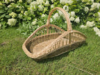 Elongated willow flower trug with a low woven base, open-weave sides and a single tall arched handle on the grass by hydrangeas