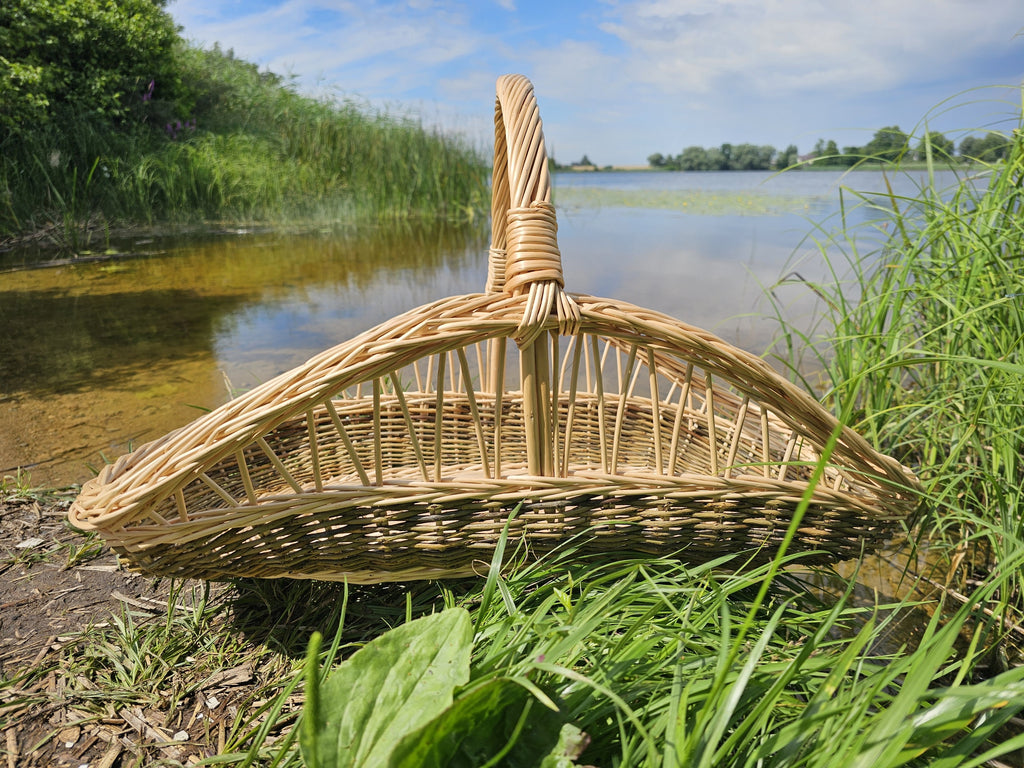 Boat-shaped willow trug with a high central arched handle on the grassy shore of a lake, the open weave running along its sides