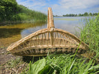 Boat-shaped willow trug with a high central arched handle on the grassy shore of a lake, the open weave running along its sides