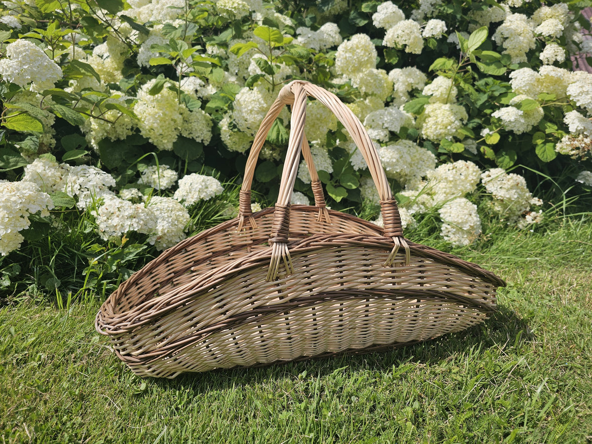 Low oval wicker trug with raised ends and two arched handles meeting at the centre, standing on grass in front of white hydrangeas