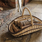 Handwoven willow garden trug used as a log basket, filled with split firewood on a stone floor beside a brick hearth.