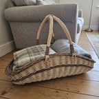 Large oval wicker trug with two handles holding folded wool throws on a wooden floor beside a grey sofa in a living room