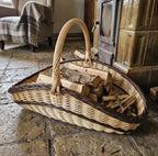 Handwoven willow trug log basket stacked with firewood on a stone floor in front of a tiled stove with a woollen throw.