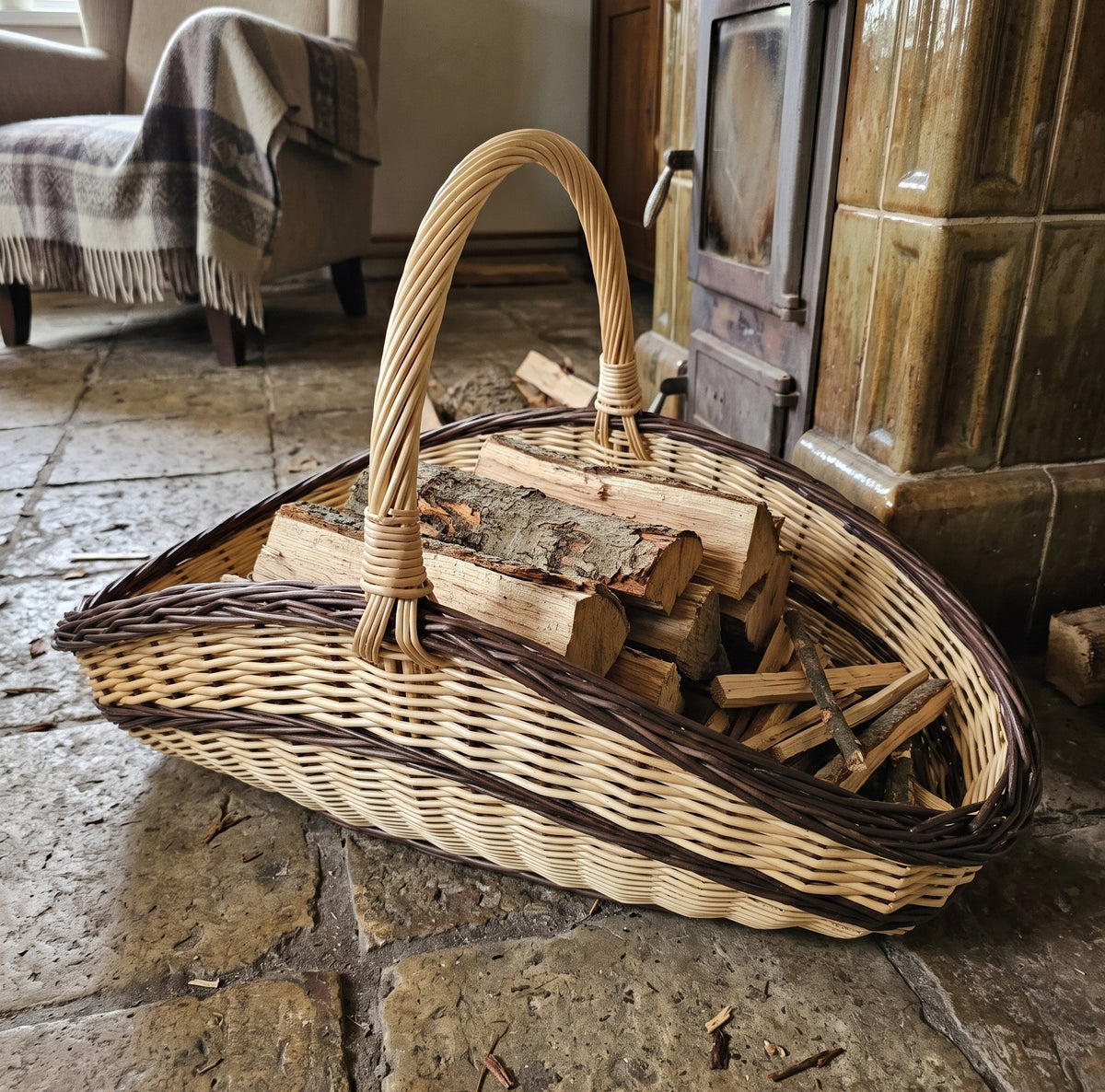 Handwoven willow trug log basket stacked with firewood on a stone floor in front of a tiled stove with a woollen throw.