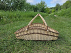 Elongated boat-shaped willow trug with two arched handles joining at the centre, standing on a grassy path in the countryside
