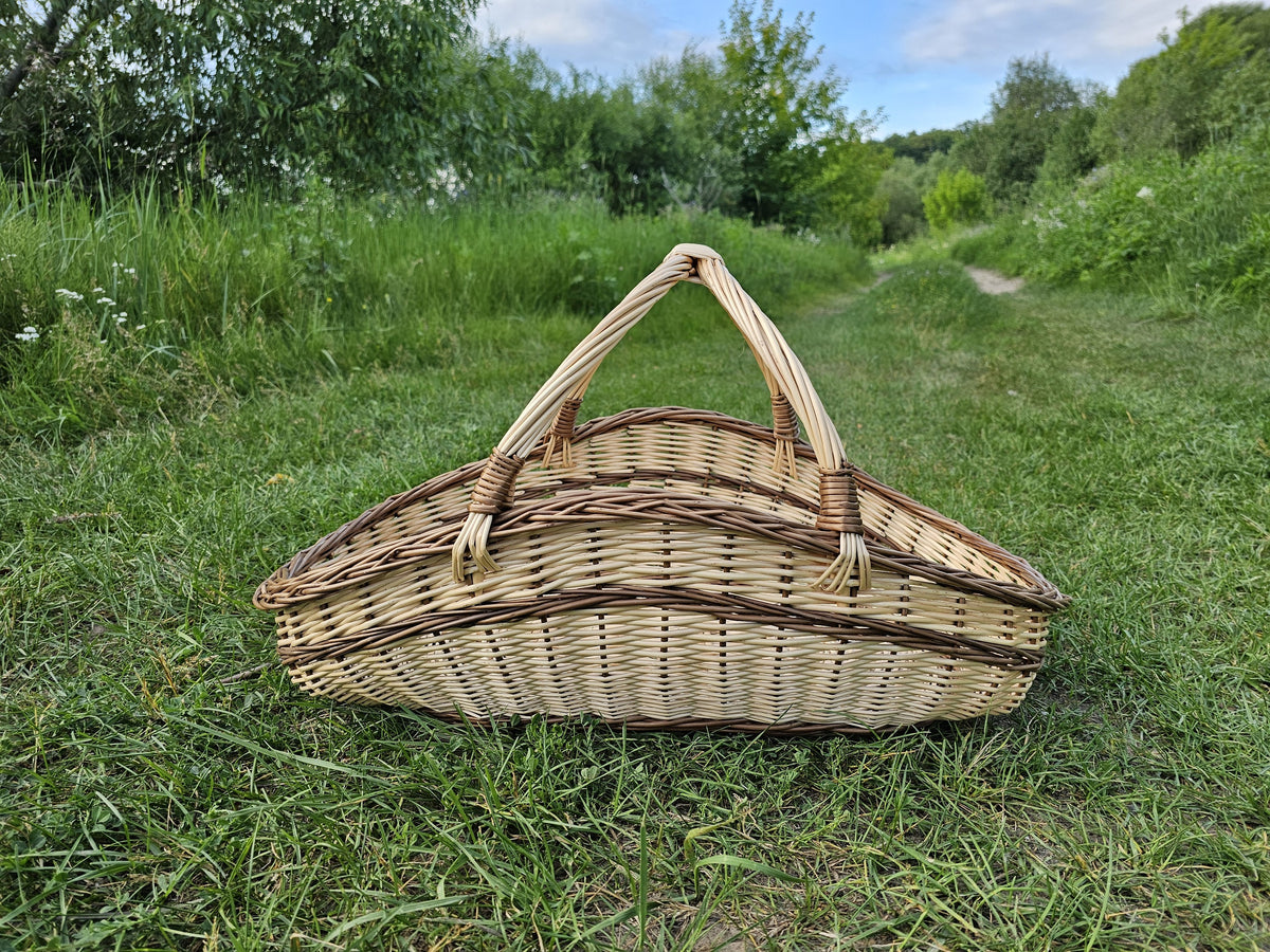 Elongated boat-shaped willow trug with two arched handles joining at the centre, standing on a grassy path in the countryside